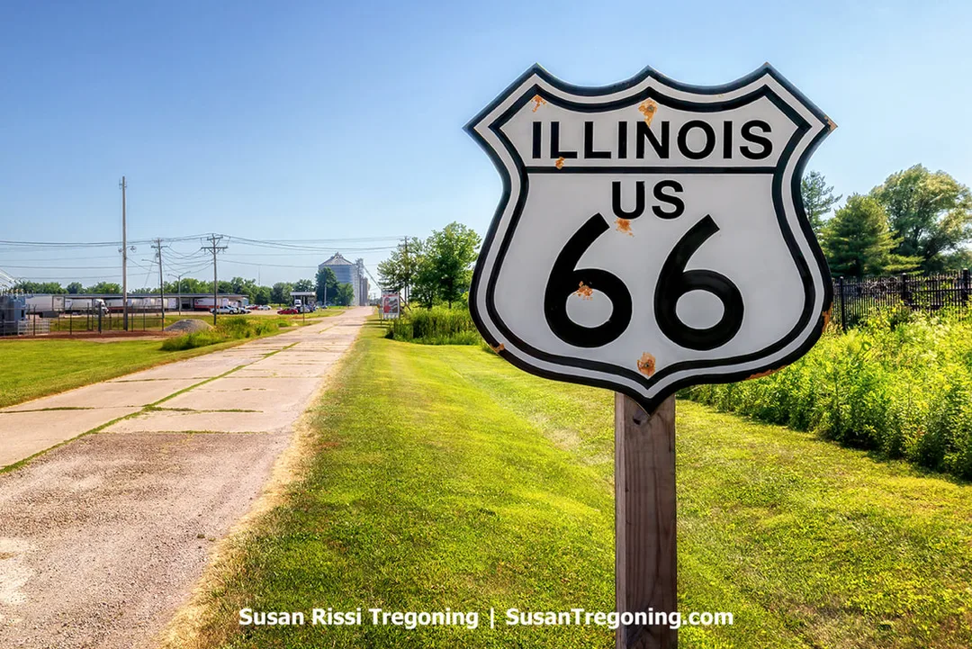 A vintage Route 66 sign stands beside a quarter‑mile stretch of original pavement from the 1926 Route 66 alignment in Atlanta, Illinois. The concrete roadway extends into the distance with grass and trees along the edges.