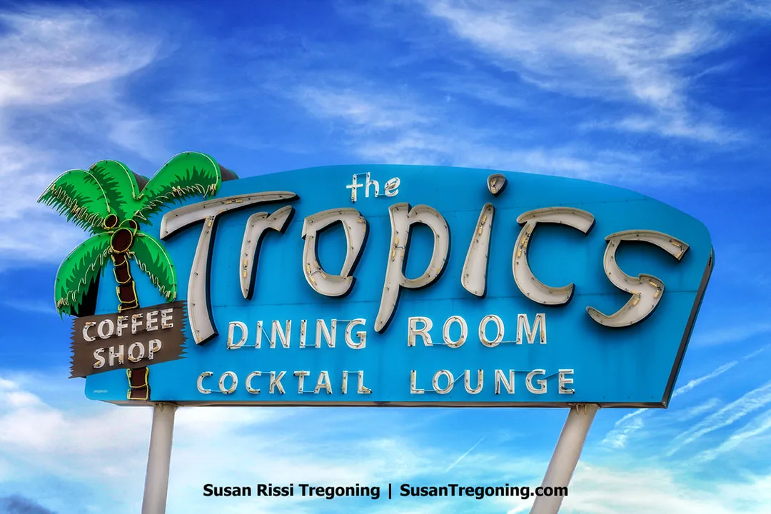 The original neon sign for The Tropics Coffee Shop, Dining Room, and Cocktail Lounge is shown in Lincoln, Illinois, along historic Route 66. The sign features multiple illuminated sections with the business name and a neon palm tree element.