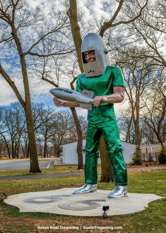 A tall fiberglass statue known as the Gemini Giant stands outdoors at its new location on Route 66 in Wilmington, Illinois. The figure wears a shiny green outfit, metallic boots, and a silver helmet, and holds a silver rocket labeled Gemini Giant. Trees, small buildings, and a partly cloudy sky appear in the background.
