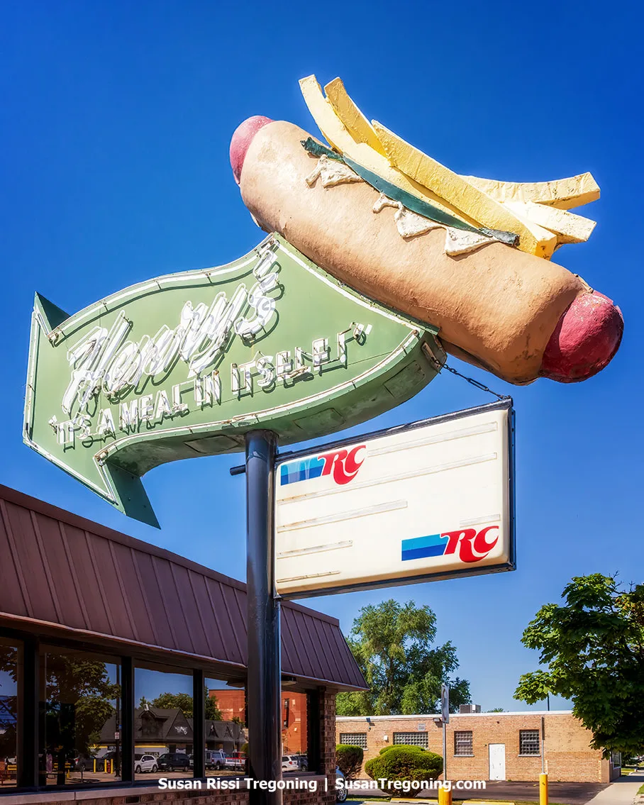 Large sculpted hot dog with fries mounted above the Henry’s Drive‑In neon sign, with a building and clear sky in the background.