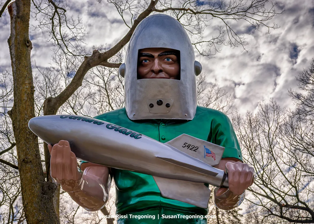 A tall fiberglass statue known as the Gemini Giant stands outdoors on Route 66 in Wilmington, Illinois. The figure wears a green shirt and a metallic helmet with a rectangular visor and holds a silver rocket marked with text and an American flag decal. Leafless trees and a cloudy sky form the background in its new location.