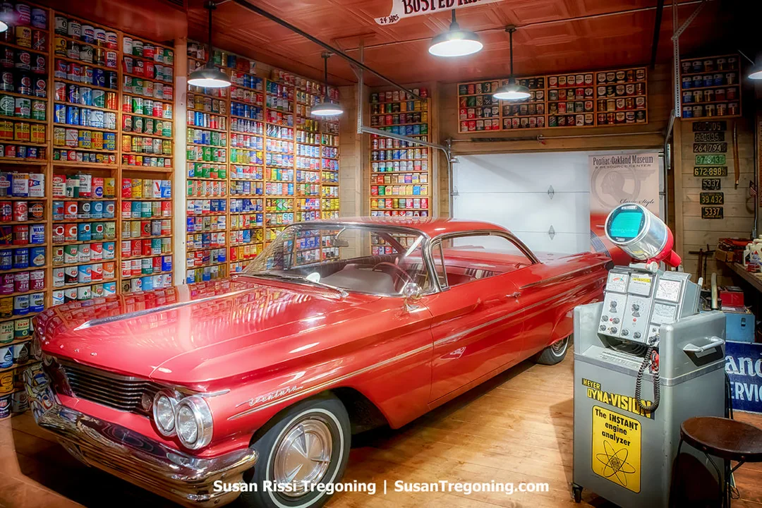 A 1960 Pontiac Ventura is parked inside a museum garage. Shelves filled with hundreds of colorful oil cans cover the walls behind the car, and vintage automotive equipment appears nearby. A banner identifies the space as part of the Pontiac Oakland Museum.
