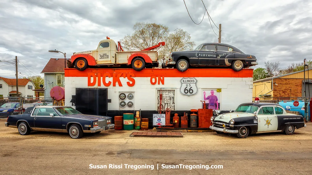  A vintage roadside building displays large painted letters reading DICK’S ON above an Illinois US 66 shield sign. Classic vehicles sit on the roof, including a red and white tow truck and a black car with California or Bust painted on the side. Three vintage cars are parked in front, along with old gas pumps, barrels, and automotive parts. The site is located on historic Route 66 in Joliet, Illinois.