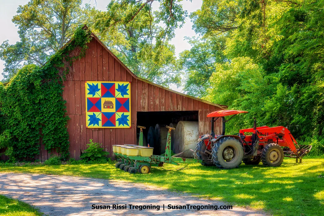 A red barn displays a large Maple Leaf barn quilt on its side, designed with gold, blue, red, and green geometric shapes. A tractor is parked in front of the barn, and trees and farm structures appear around the scene.