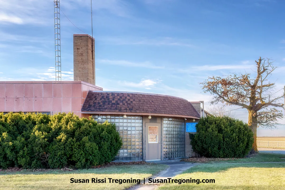 A small mid‑century building stands along a rural roadside. It features glass‑block walls, a curved shingled roof, and a pink‑tiled rectangular section. A tall brick tower rises behind the structure with a metal antenna beside it. Trimmed shrubs, a tree, and an open field appear around the building under a clear sky. This building once served as the Illinois State Police Headquarters on Route 66 just south of Pontiac.