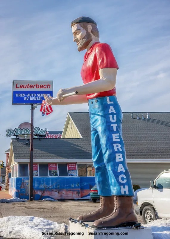 The 25‑foot‑tall Lauterbach Giant stands on the 1926–1930 alignment of Route 66 in Springfield, Illinois. The fiberglass figure is positioned in front of Lauterbach Tire & Auto Service and holds an American flag. Surrounding pavement, signage, and roadside elements are visible.