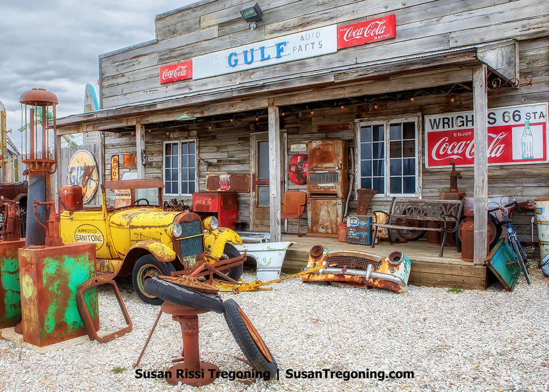 The natural‑wood Ghost Gas Station façade at Motorheads Bar, Grill, and Museum in Springfield, Illinois, is shown outdoors. The display includes vintage gas pumps, signs, and classic cars in their original rusty condition, with some pieces dating back to the 1920s.