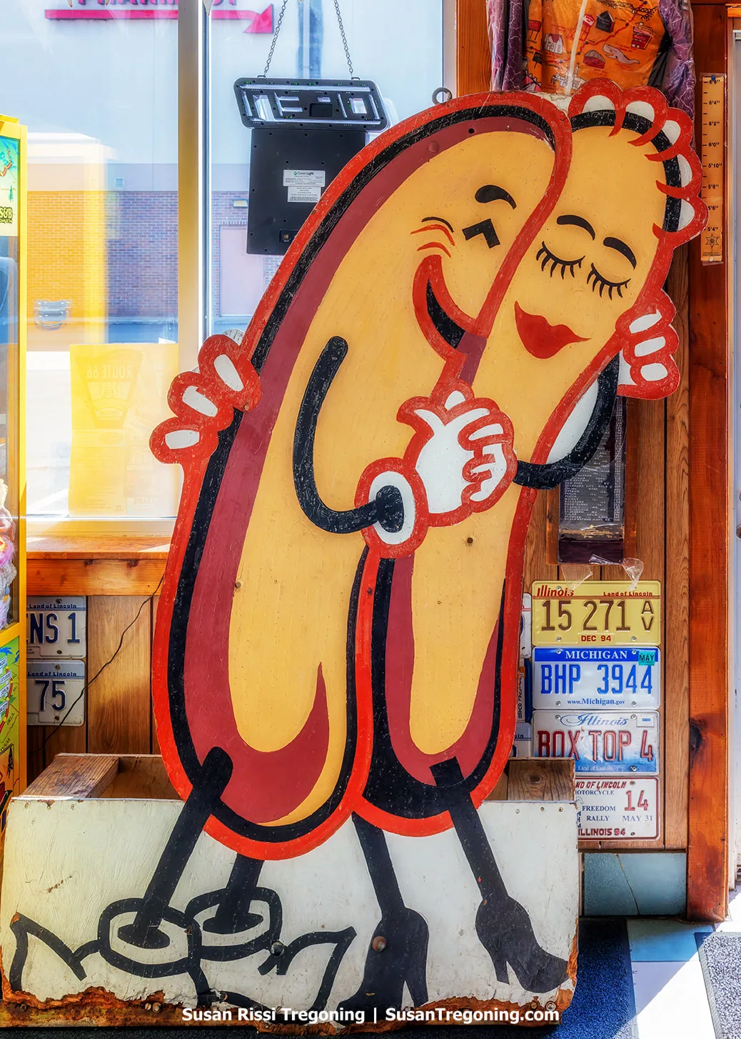 The original Cozy Dog sign is displayed inside the Cozy Dog Drive‑In on Route 66 in Springfield, Illinois. The artwork shows a male and female Cozy Dog character hugging each other, illustrated in yellow with red and black outlines.