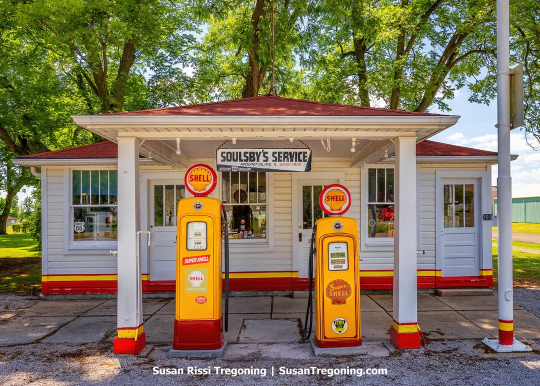 The 1926 Soulsby Service Station in Illinois is shown from the exterior. The white building with gold and red Shell branding has old Shell tanks positioned under the canopy.