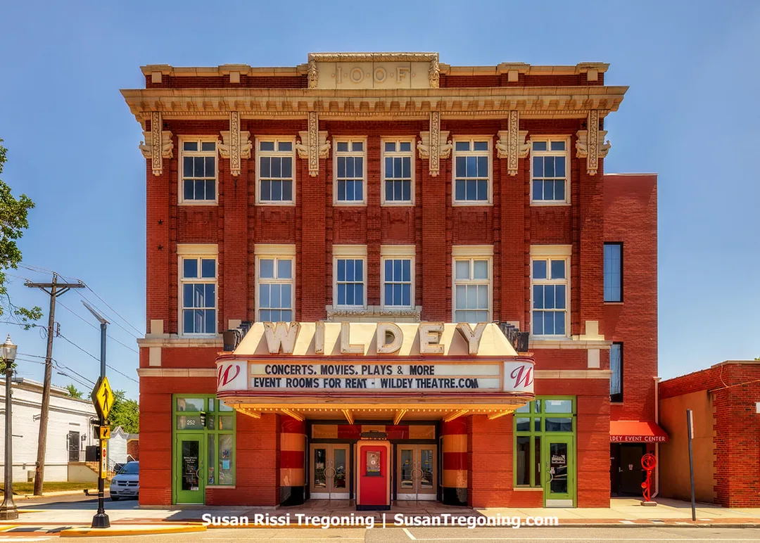 The entrance of the historic Wildey Theatre in Edwardsville, Illinois, is shown with an Art Deco ticket booth positioned between double doors. A large marquee extends above the entrance with neon letters spelling out Wildey on the front and sides.