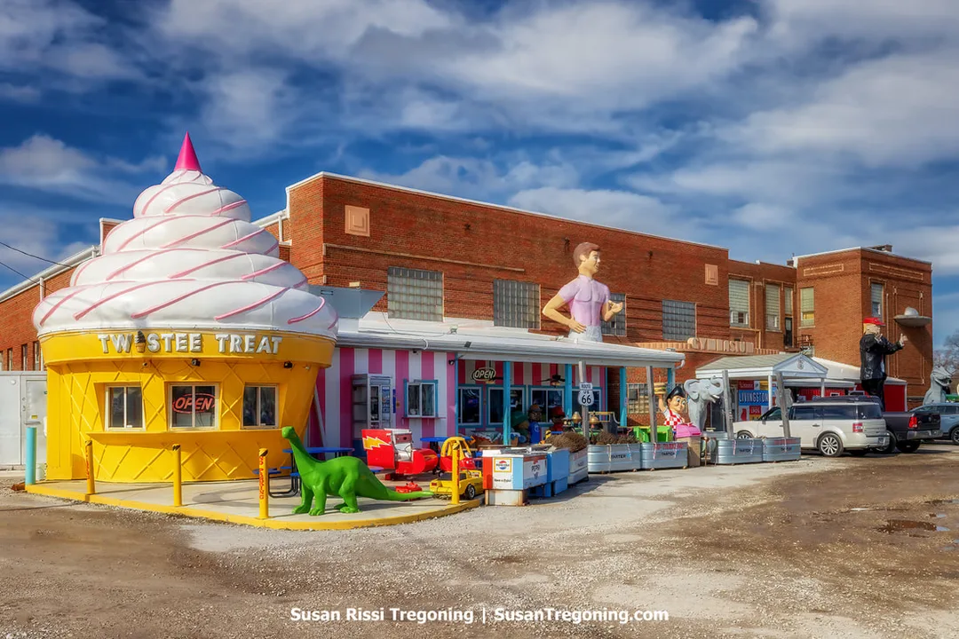 The exterior of the Pink Elephant Antique Mall in Livingston, Illinois, is shown along historic Route 66. The building is the former 1929 Livingston High School, with a large multi‑story brick façade and rows of windows. A Twistee Treat ice‑cream‑cone‑shaped diner stands on the property, along with several giant outdoor statues. Additional 1950s‑style retro elements are visible around the grounds.