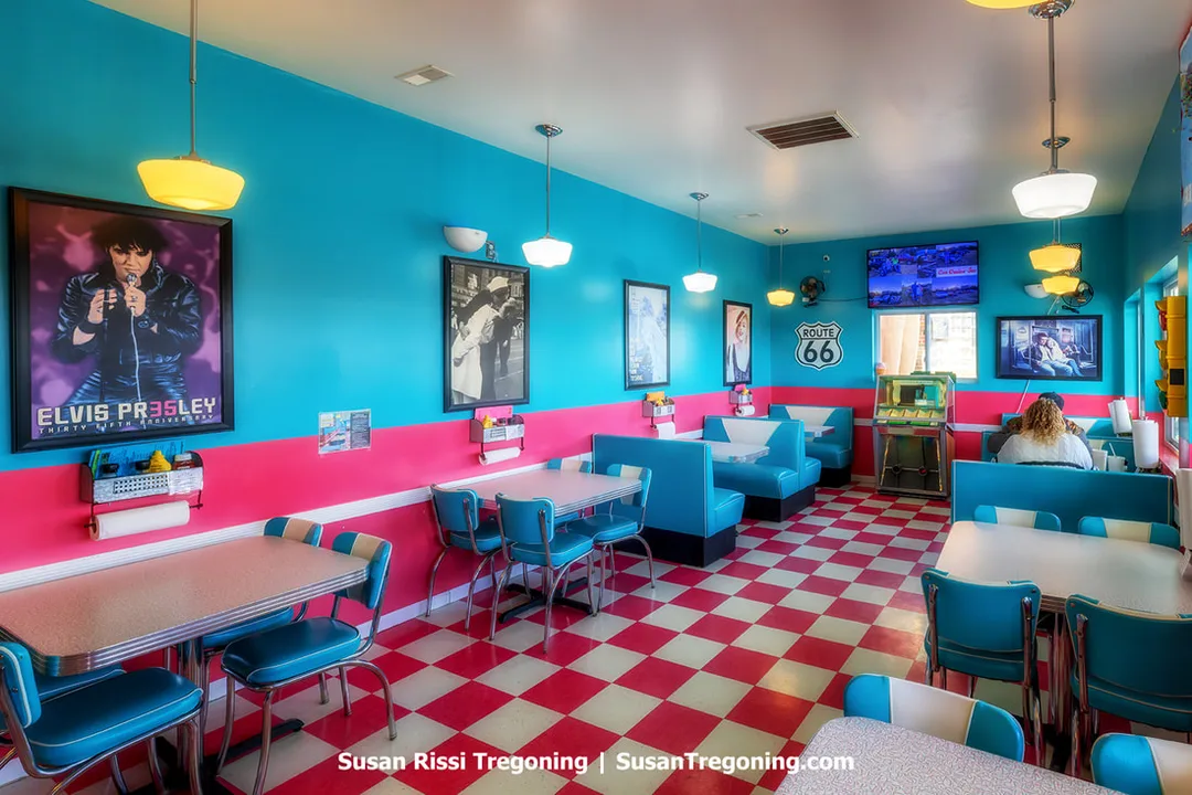 The interior of the Twistee Treats Diner shows a retro 1950s‑style space inside an original ice‑cream‑cone‑shaped building. The dining area has hot pink and white checked tile and turquoise walls, with images of Elvis Presley and other 1950s icons displayed on the walls.