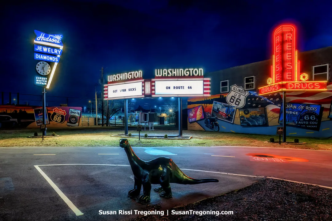 It’s Electric – Neon Sign Park in Granite City, Illinois, is shown at night with recreated neon signs for Hudson’s Jewelry and Diamonds, the Washington Drive‑In, and Reese’s Drugs. A Sinclair Dino figure stands in the parking lot. The site is one of the newest Route 66 roadside attractions in the state.