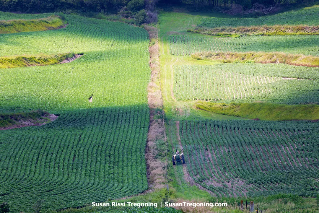 A bird’s‑eye view of a farmer riding a tractor along a fence line in the Loess Hills. The tractor follows the contour of a steep, wind‑formed hillside while the surrounding field shows the uneven, rolling terrain characteristic of the region.