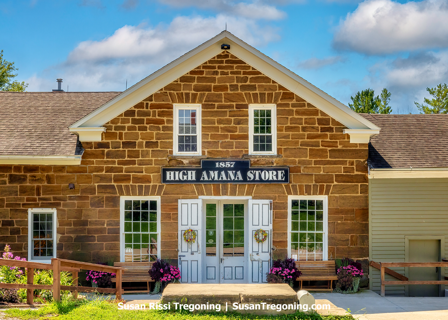 Historic 1857 High Amana Store building with a brown‑stone exterior, white trim, and a gabled roof. The front façade has three upper windows, three lower windows, and a centered double‑door entrance decorated with wreaths. A black sign above the door reads “1857 HIGH AMANA STORE.” Benches, flower arrangements, and a wooden ramp sit at the entrance. Greenery surrounds the well‑maintained building under a bright blue sky with scattered clouds.