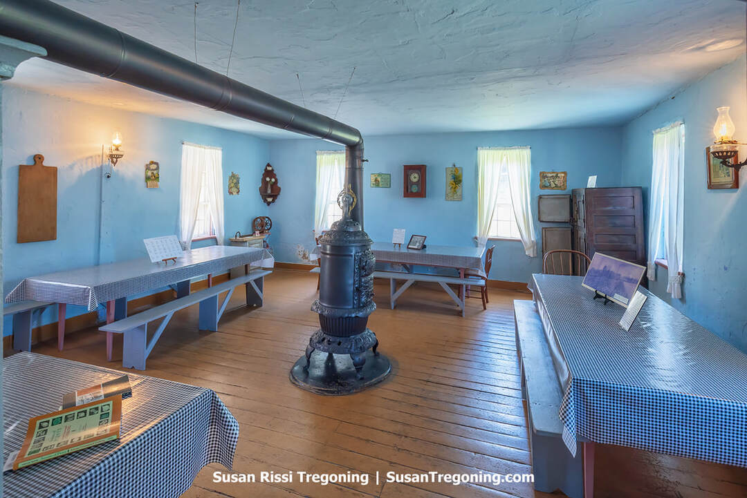 Inside the Ruedy Communal Kitchen dining area, long wooden tables are covered with navy‑and‑white checked tablecloths. The walls are painted Colony Blue, a traditional Amana color, and the room is arranged with simple wooden benches and neatly set tables that reflect the historic communal dining traditions of the Amana Colonies.