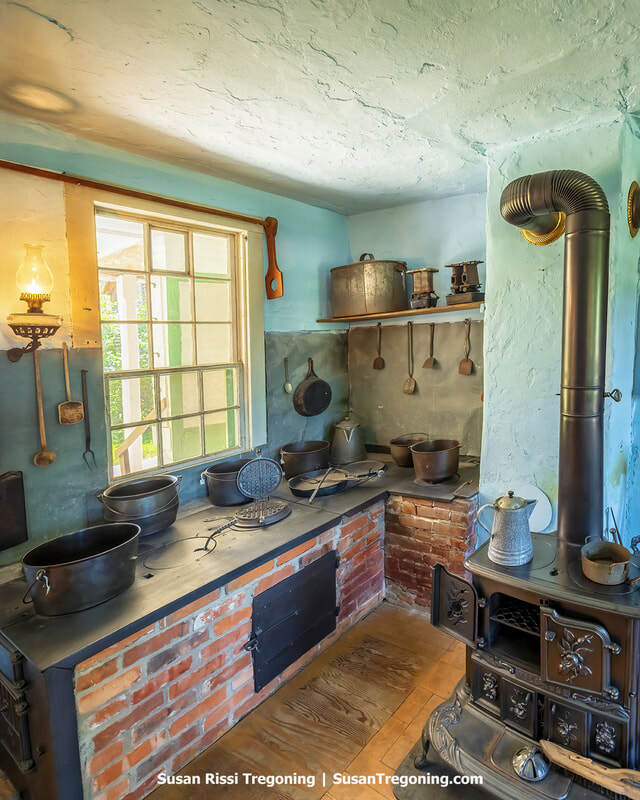 Inside the Ruedy Communal Kitchen cooking area, with worktables, shelves, and historic food‑preparation tools arranged in a preserved 19th‑century layout. The space shows the utilitarian setup used for preparing communal meals in the Amana Colonies.