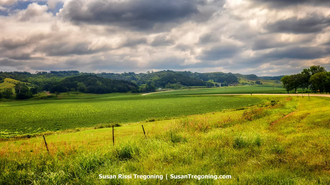 A roadside view while driving the Byway Spine on Iowa’s Loess Hills National Scenic Byway. The paved road curves through open farm fields, with the rolling, loess‑formed hills visible in the distance.