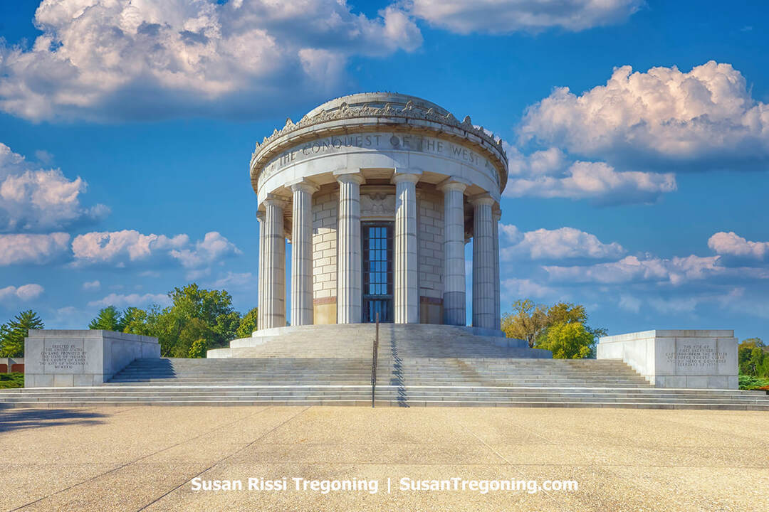 The circular, columned George Rogers Clark Memorial stands at George Rogers Clark National Historical Park in Vincennes, Indiana. The neoclassical structure features a domed roof and tall stone columns, with engraved inscriptions on nearby stone blocks. Trees frame the scene under a partly cloudy sky.