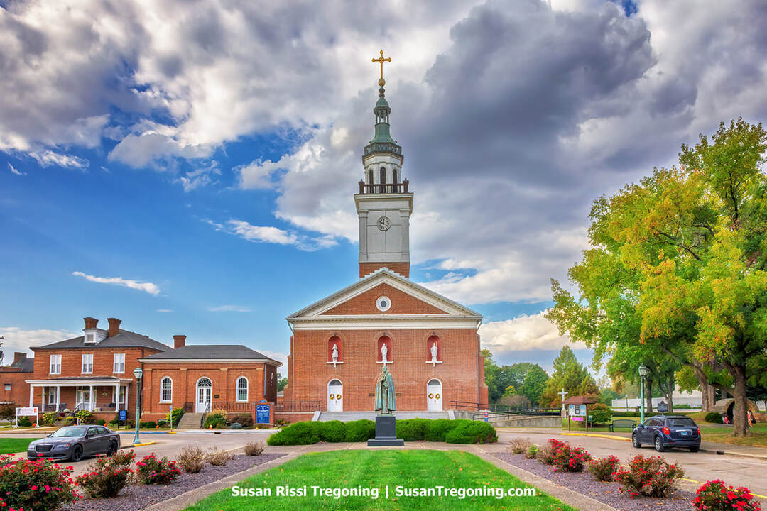 The historic Basilica of St. Francis Xavier, part of the Old Cathedral Complex in Vincennes, Indiana, stands along a town street. The brick church features a tall steeple topped by a gold cross, with three arched façade niches holding statues and a larger statue on a pedestal in the landscaped garden at the entrance. Nearby brick buildings and parked cars line the street beneath a partly cloudy sky.