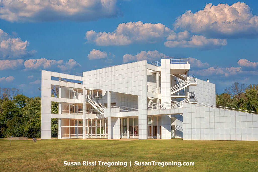 Richard Meier’s Atheneum rises in crisp white geometric planes, with stacked exterior staircases and open terraces creating a sculptural, multi‑level form. Large windows punctuate the minimalist façade, and the building stands against a backdrop of trees and sky above a wide, grassy lawn, emphasizing its clean lines and modernist symmetry.