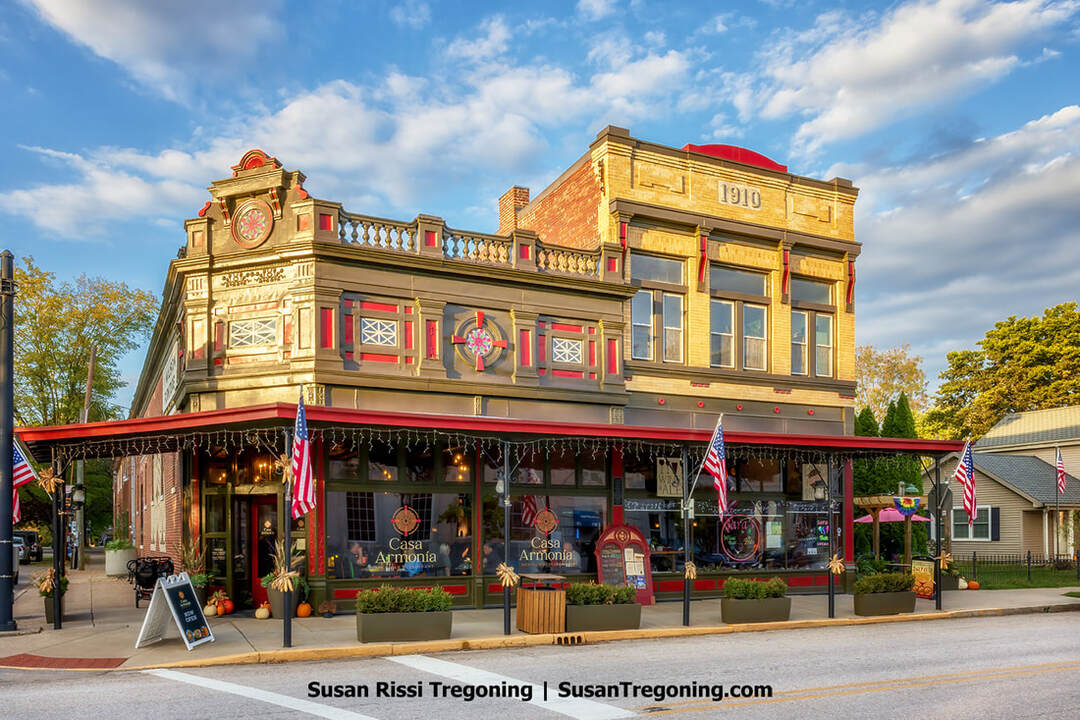 A two‑story historic commercial building with ornate classical detailing stands along a sunny streetscape. The right section, marked with the year 1910, features tall windows framed by decorative moldings and a balustrade along the roofline. The left section has red accents and a circular emblem near the top. On the ground floor, Casa Armonia occupies the storefront, with pumpkins, planters, American flags, and string lights creating a welcoming entrance. A sandwich‑board sign and additional potted plants line the sidewalk beneath a partly cloudy sky.