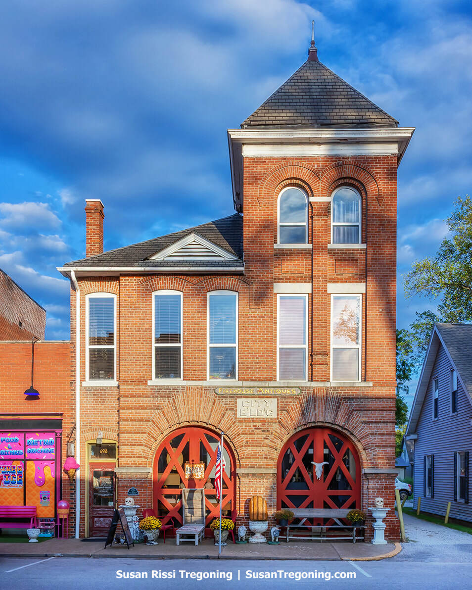 A historic red‑brick firehouse with a tall, steep‑roofed tower stands along a sunny streetscape. The arched red doors are marked “City Bldg,” and the façade features decorative brickwork and arched windows. The building now houses an antique shop, with benches, flowerpots, flags, and vintage items arranged outside. A brightly colored storefront sits just beyond it under a blue sky with scattered clouds.