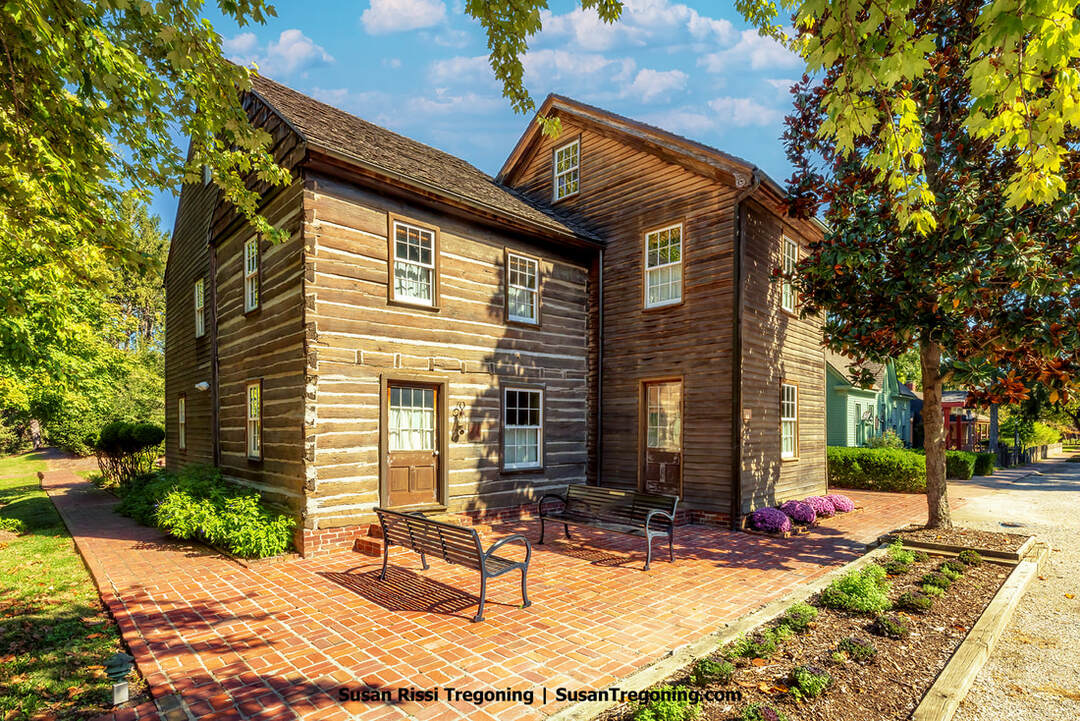 A two‑story log building with horizontal timbers and white chinking stands on a red brick patio, its white‑trimmed windows and twin doors facing two black metal benches. Trees and shrubs surround the structure, and neighboring houses are visible beyond the garden on a bright, sunny day.