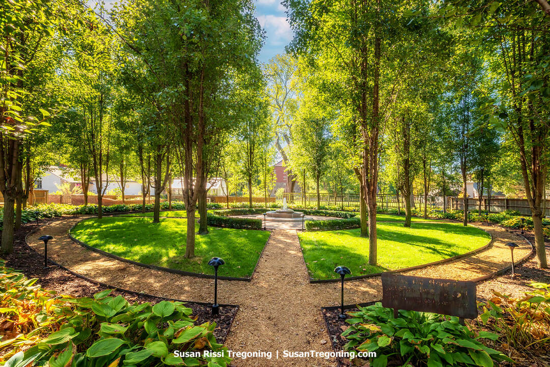 The Carol Owen Coleman Memorial Garden features a circular lawn and gravel paths arranged in a symmetrical design, leading toward a central stone monument framed by evenly spaced, column‑straight trees. Low shrubs, flower beds, and small pathway lights border the walkways, creating a serene, formal garden setting under soft autumn color.