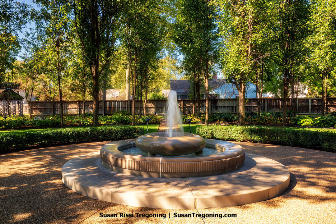 A circular stone fountain sprays water upward from a raised central basin, set within a paved courtyard bordered by neatly trimmed hedges. Tall trees cast dappled shade around the Fountain of Life, with a wooden fence and glimpses of nearby homes visible through the foliage, creating a quiet, residential garden setting.