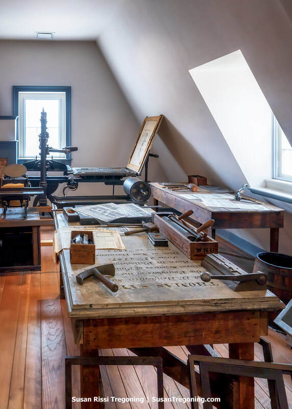 A historic print shop is set up in an attic-like room with angled ceilings, wooden floors, and two windows filling the space with natural light. A platen press stands at the back, while worktables in the foreground hold typesetting trays, printed sheets, and blocks of type. Tools and materials are arranged neatly across the wooden surfaces, creating an authentic early printing‑room atmosphere.