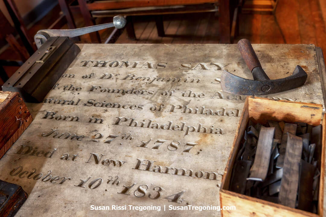 A close‑up view of Thomas Say’s original tablet‑style tombstone shows the engraved stone resting on a wooden workbench, its surface surrounded by traditional typesetting tools. Metal letter stamps, a wooden box of type, and other printing implements sit nearby, reflecting how the stone was later repurposed as a work surface in the Slater Printing Office on the third floor of Harmonist Community House No. 2 in New Harmony, Indiana.