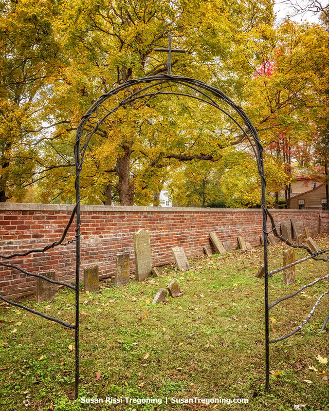A wrought‑iron archway topped with a cross frames the view into an old cemetery enclosed by a red brick wall. Weathered gravestones lean at varied angles beneath tall trees filled with bright yellow and orange autumn leaves. A house is partially visible through the foliage in the background, and the scene is quiet and reflective.