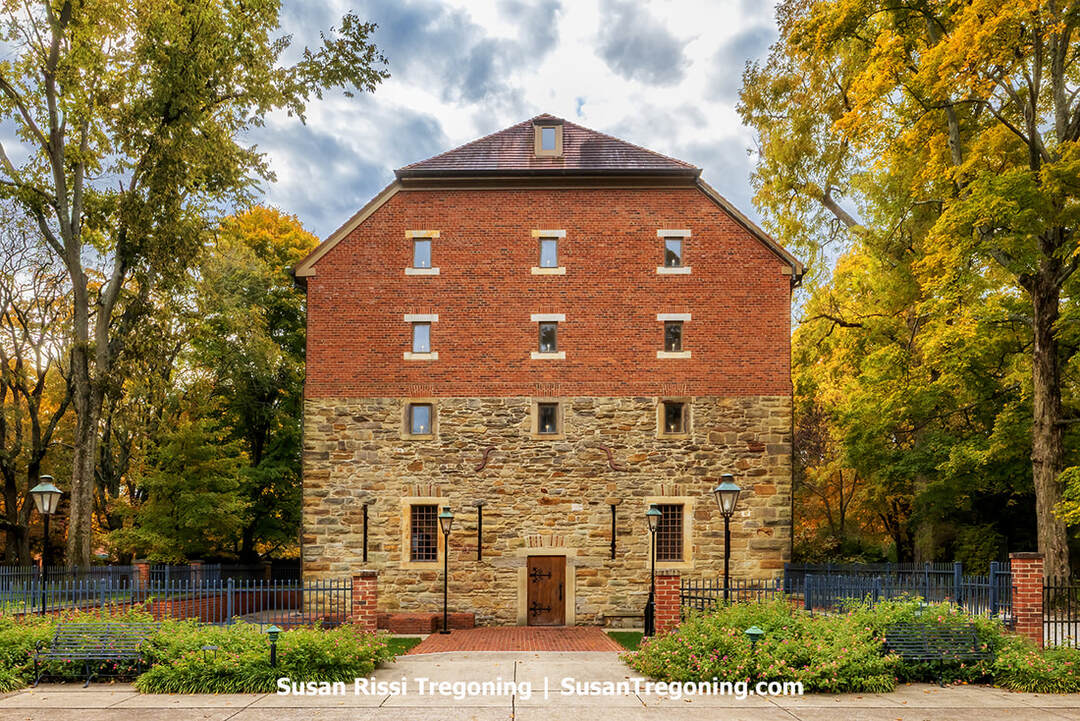 A two‑story granary built of rough stone on the lower level and red brick above stands among autumn trees, its symmetrical façade centered on a simple door and evenly spaced windows. Vintage-style streetlamps and benches sit in the foreground, and fallen leaves scatter across the quiet, park‑like setting surrounding the historic Rapp‑Owen Granary.