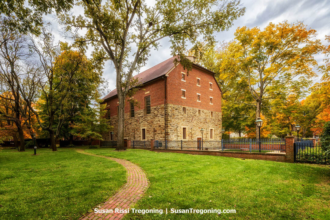 A two‑story brick and stone granary in New Harmony, Indiana, stands behind a black iron fence with brick pillars and vintage-style lamps. The building’s stone lower level and red‑brick upper walls are framed by mature trees with autumn foliage. A curved brick walkway crosses a neatly mowed lawn in the foreground, leading toward the historic structure.