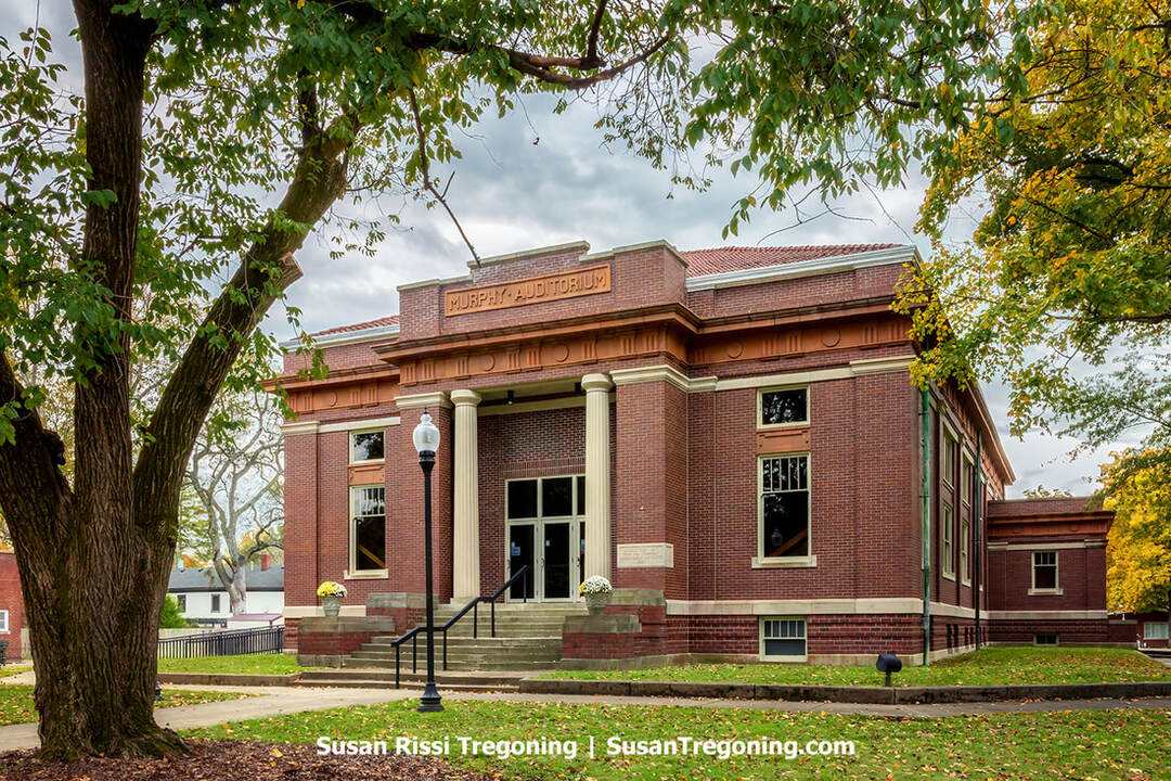 Murphy Auditorium, a stately red‑brick building with a classical façade, features a columned entrance framed by decorative stonework and an engraved nameplate above the doors. A concrete walkway leads to the front steps, flanked by a lamppost and flower pots, while mature trees with autumn foliage surround the structure and soften its formal architectural lines.