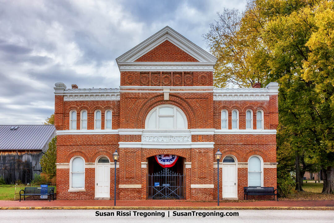 A historic red‑brick opera house stands beneath an overcast sky, its arched central doorway marked by the engraved words “Opera House.” White trim highlights the tall arched windows, and patriotic bunting hangs above the entrance. Two vintage-style streetlamps flank the doorway, benches sit on either side, and autumn trees with orange and yellow leaves frame the building.
