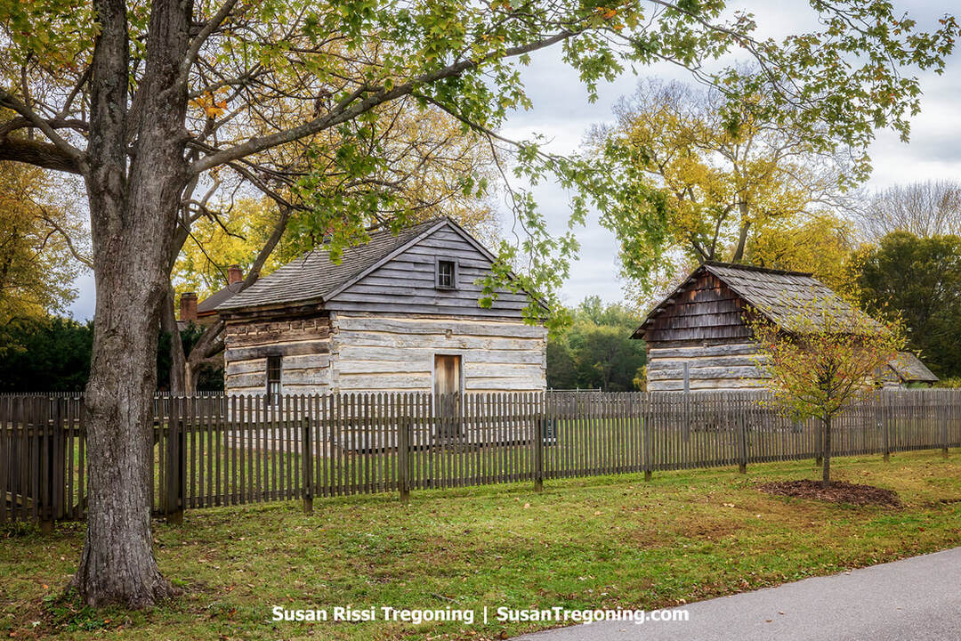 Two log blockhouses stand inside a low wooden picket fence, their hand‑hewn horizontal timbers and simple shingled roofs set against mature trees with autumn foliage. A grassy lawn surrounds the structures, and soft fall light highlights the texture of the historic cabins.