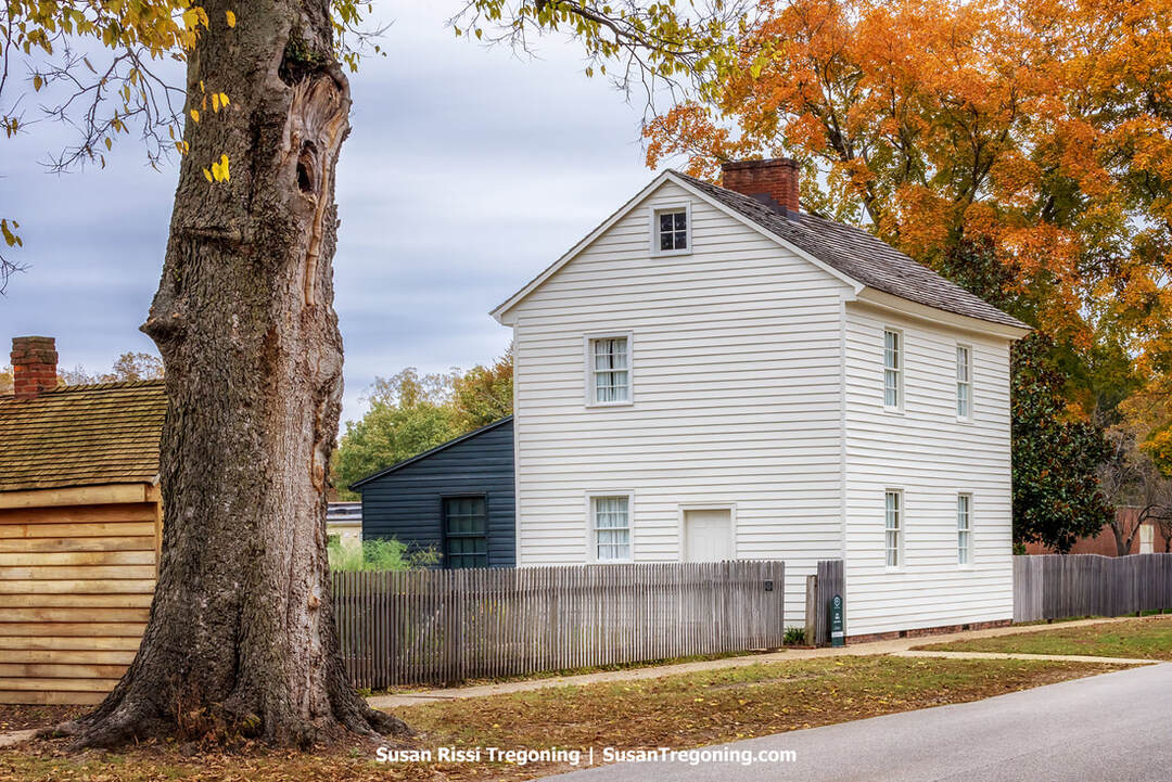 A white two‑story frame house with a gabled roof stands behind a wooden picket fence on a quiet street. A large tree trunk fills the left foreground, and a smaller outbuilding with a shingled roof sits beside the house. Autumn trees with yellow and orange foliage rise behind the buildings, and the overcast light softens the historic scene.
