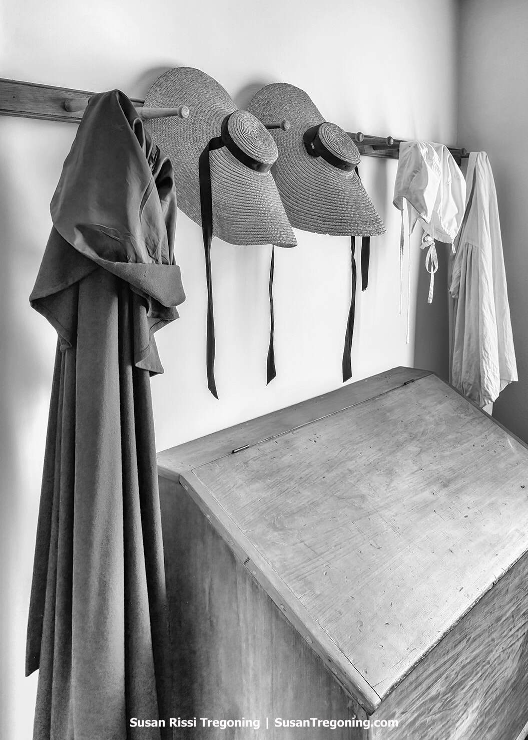 Alt text:
A row of Harmonist garments hangs from wooden pegs above a slant‑lidded wooden chest: a long cloak, two wide‑brimmed straw hats with dark ribbons, a bonnet, and a light-colored dress or apron. The scene is rendered in black and white, emphasizing the textures of the clothing and the simple furnishings inside the historic Lenz House.