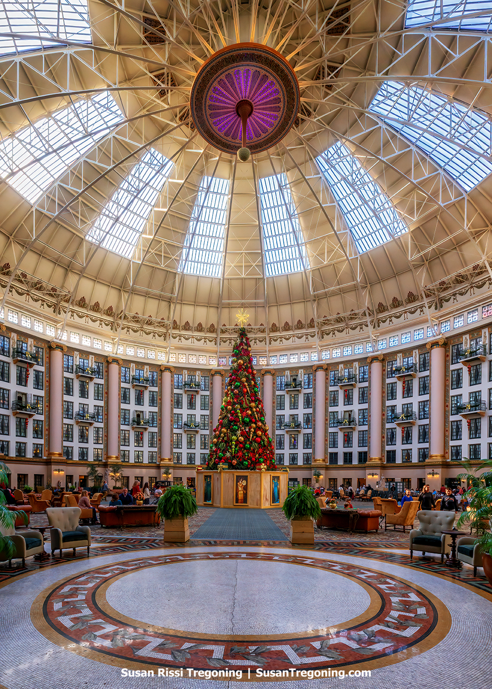 A soaring hotel atrium with a glass‑paned domed ceiling rises above a tall, fully decorated Christmas tree set at the center of a circular mosaic floor. Multiple stories of balconies, columns, and arched windows frame the space, with seating areas, plants, and people gathered throughout the lobby. The festive tree stands on a wooden base displaying framed artwork.