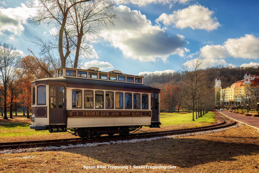 A vintage trolley labeled “French Lick and West Baden Railway” travels along a curved track through a park-like setting. Leafless trees line the path, and a large yellow historic hotel with red roofs and towers rises in the background under partly cloudy sunlight.