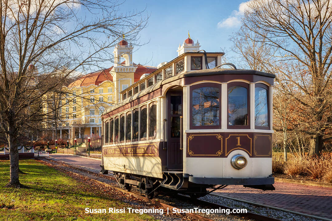 A vintage red and cream streetcar numbered 1 sits on a track beside a brick drive. Behind it stands a large yellow historic hotel with red roofs, white trim, and multiple towers. Leafless trees frame the scene in warm sunlight.
