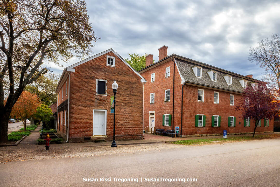 Community House No. 2 in New Harmony, Indiana, stands as a large three‑story brick structure with green shutters and dormer windows, set along a quiet tree‑lined street. A smaller brick building sits beside it, and a vintage-style streetlamp, benches, and autumn foliage contribute to the historic, village‑like atmosphere.
