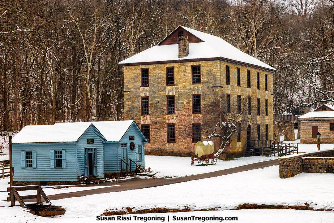 The three‑story 1817 limestone grist mill at Spring Mill State Park dominates the snowy pioneer‑village scene, its tall stone walls rising behind two small blue wooden shops in the foreground. Snow blankets the ground and rooftops, a covered wagon sits near the mill, and leafless trees fill the winter background.