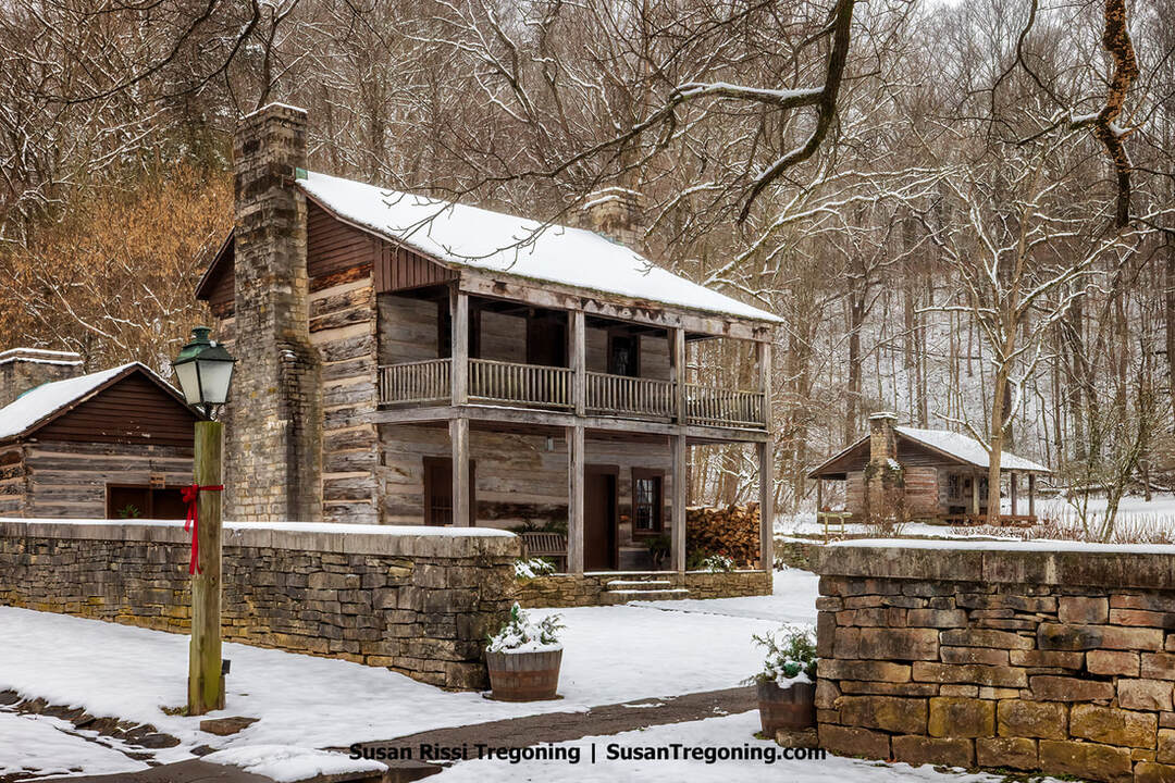 The Upper Residence in the Pioneer Village at Spring Mill State Park is shown in winter, with snow covering the log cabin’s roof, balcony, and surrounding grounds. The two‑story cabin features a stone chimney and sits beside smaller wooden outbuildings enclosed by a low stone wall. A vintage-style lamppost with a red ribbon stands in the foreground, and leafless snow‑dusted trees fill the background.