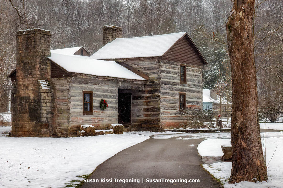 The Weaver Cabin at the entrance to Spring Mill State Park’s Pioneer Village is shown in winter, with snow covering its log walls, steep roof, and stone chimney. A Christmas wreath hangs beside the doorway, and a paved path leads to the entrance. Bare trees rise behind the cabin, and a large tree trunk frames the snowy scene on the right.
