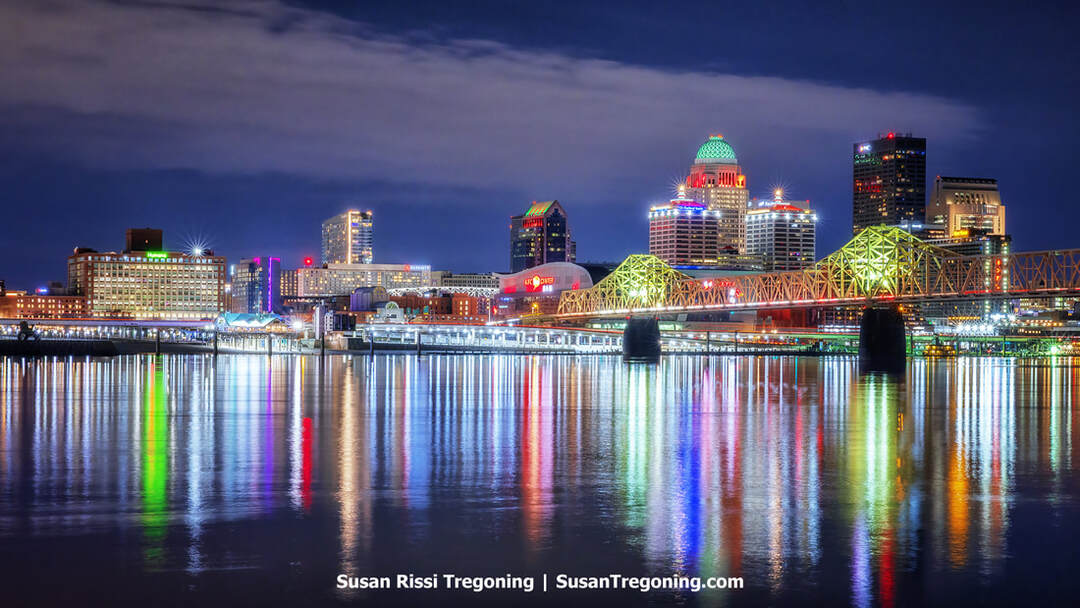 The Louisville skyline glows at night with illuminated high‑rise buildings reflecting across the dark Ohio River. The George Rogers Clark Memorial Bridge stretches across the water at the left, its lights forming a bright line leading toward the city’s tallest towers against the night sky.
