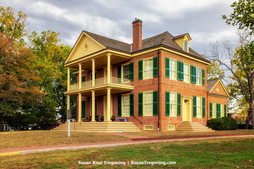 Grouseland, the brick Federal‑style home of William Henry Harrison in Vincennes, Indiana, features green shutters, yellow trim, and a two‑story columned portico. A historical marker stands near the front steps, with mature trees and a manicured lawn surrounding the house under a partly cloudy sky.