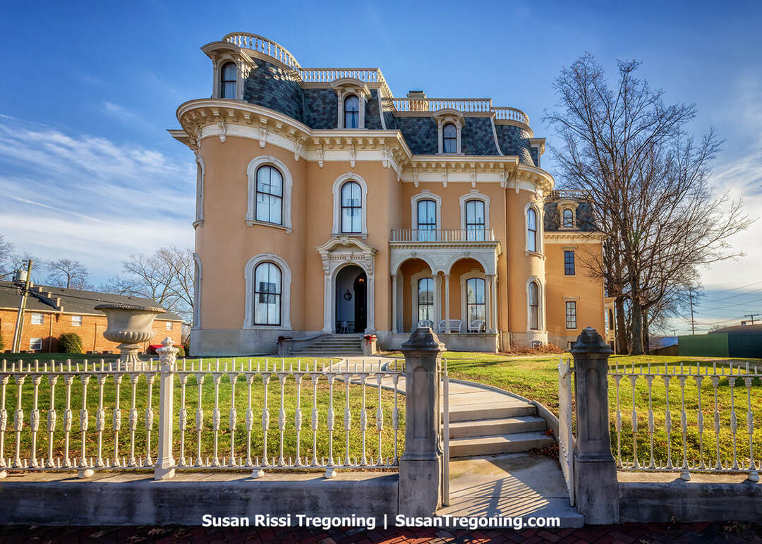 A large Victorian mansion with ornate architectural detailing rises behind a wrought‑iron fence. The multi‑story brick home features tall windows, decorative trim, and a prominent tower, with bare trees framing the historic structure.
